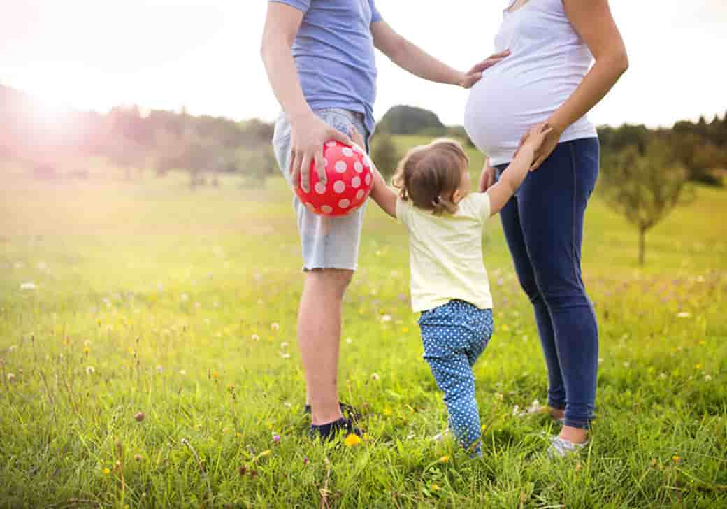 A father, pregnant mother, and child playing with a ball in the grass.