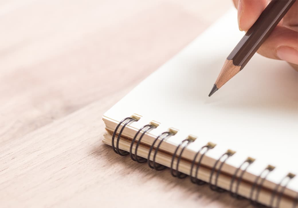 A hand writing with a pencil in a spiral notebook.