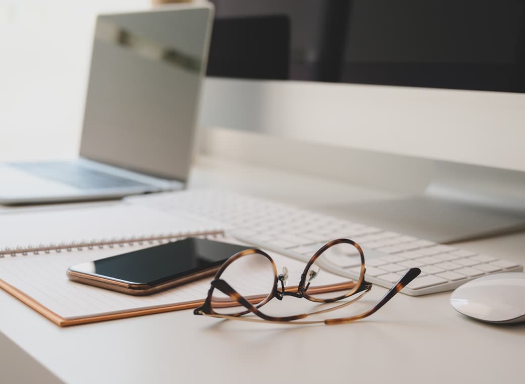 A pair of reading glasses set on a desktop with a computer and mobile phone.