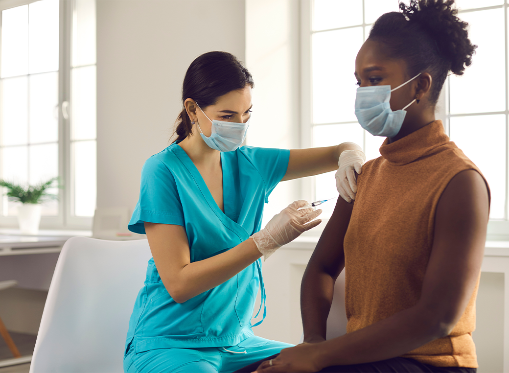 Young African American lady sitting at doctors office and getting modern Covid 19 shot. Nurse in medical face mask and gloves holding syringe and giving female patient flu vaccine injection in arm