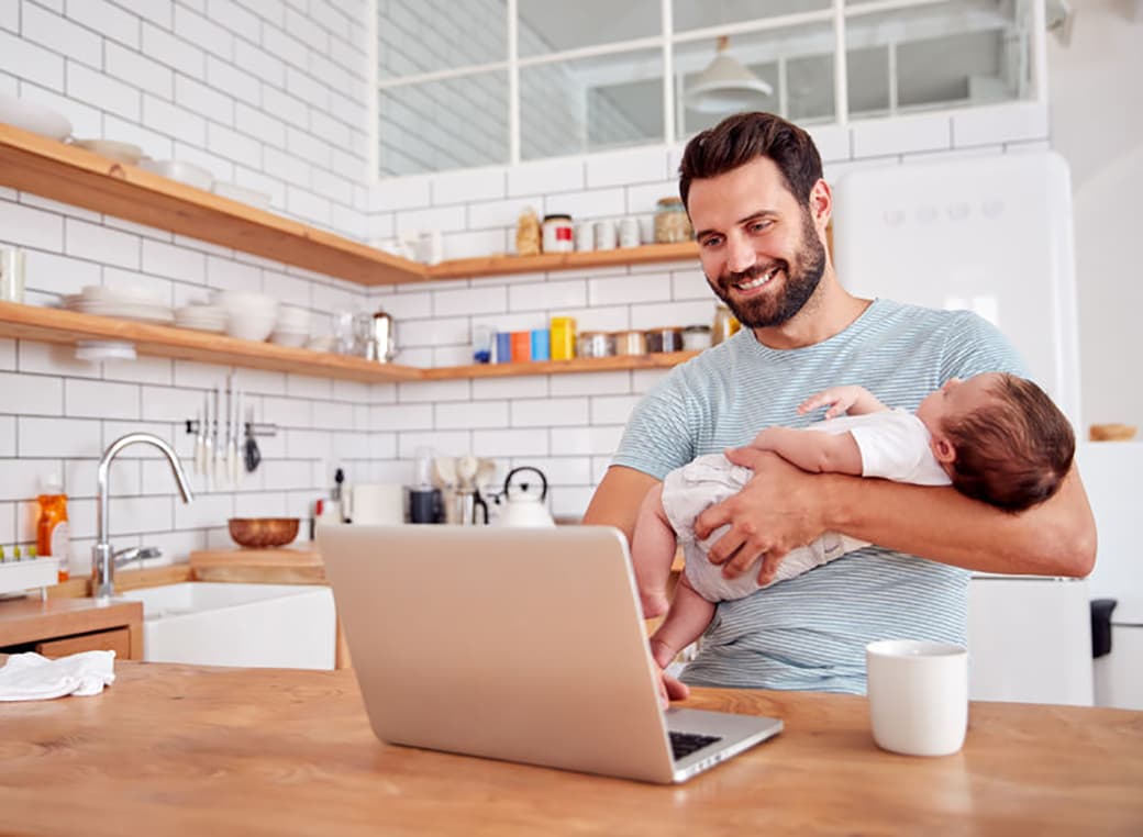 Multi-tasking father holds sleeping baby and works on laptop computer In kitchen.