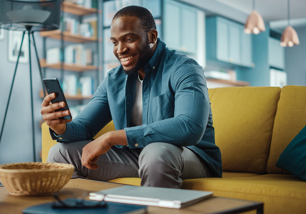 A smiling man holding and looking at a smart phone sitting on a yellow couch.