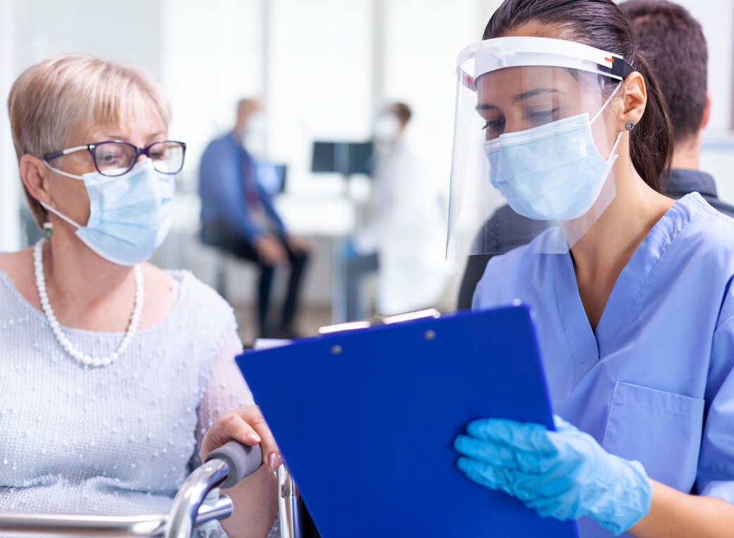 Medical staff wearing face visor in hospital hallway talking to senior woman wearing face mask.